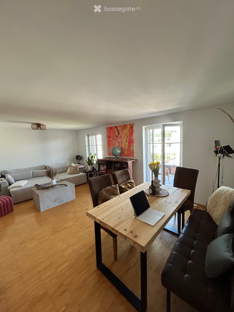 A modern living room with a wooden floor, a grey sofa, a coffee table, a laptop on a wooden table, and a leather armchair. There is a large window with a view of the balcony and a potted plant on the windowsill.