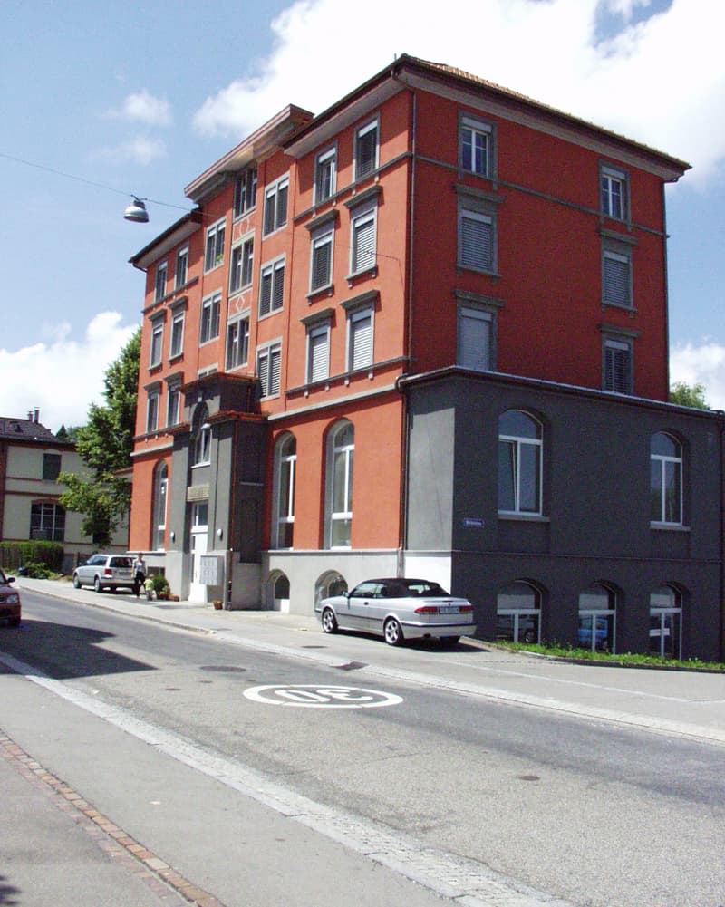 A multi-story red brick building with white-framed windows and a grey lower section, located on a street with parked cars and a pedestrian crossing sign.