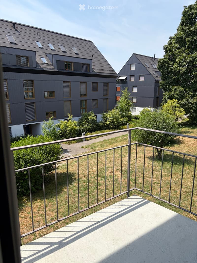 Balcony overlooking a grassy area with modern buildings in the background, featuring a metal railing and concrete floor.