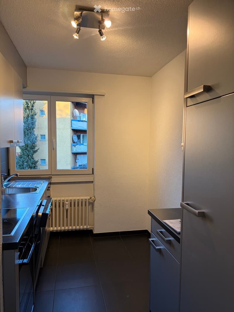 Modern kitchen with dark tiled floor, light-colored walls, stainless steel sink, oven, radiator, and cabinets.
