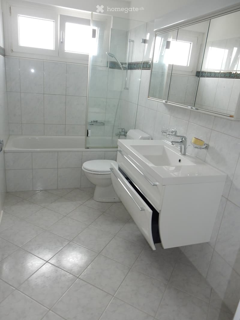 Modern bathroom with white tiles, a bathtub, a toilet, a sink with a mirror, and a glass-enclosed shower area.