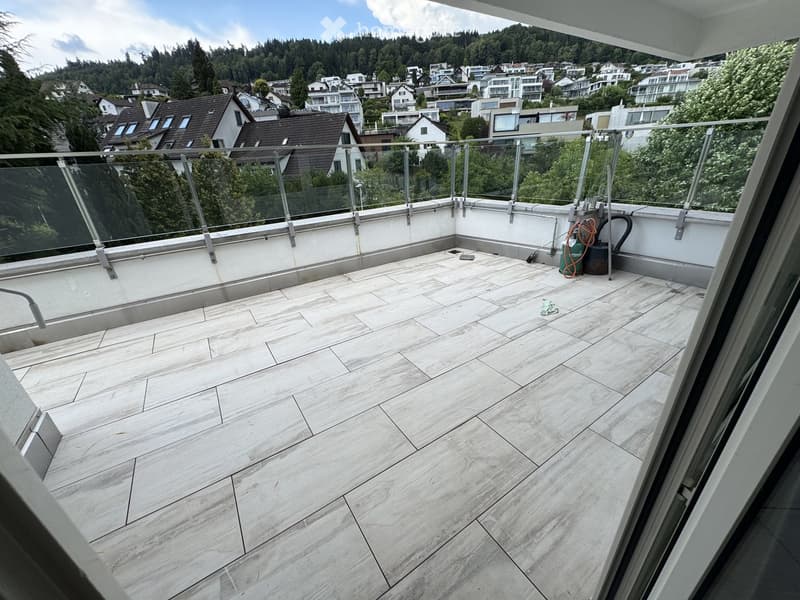 Modern balcony with marble tiles, glass railing, and scenic view of a town and forested hills.