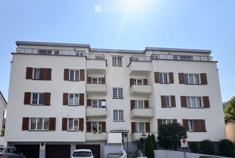 Four-story residential building with white facade, multiple balconies, and brown shutters. Features a garage area with parked cars and a small garden area with trees.
