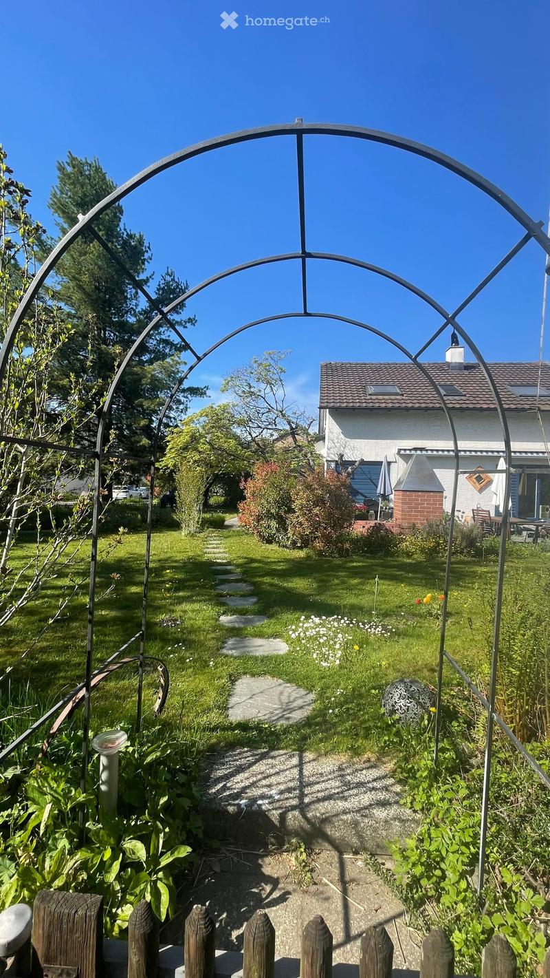 Garden with a metal archway, stone pathway, lush greenery, and a house in the background.