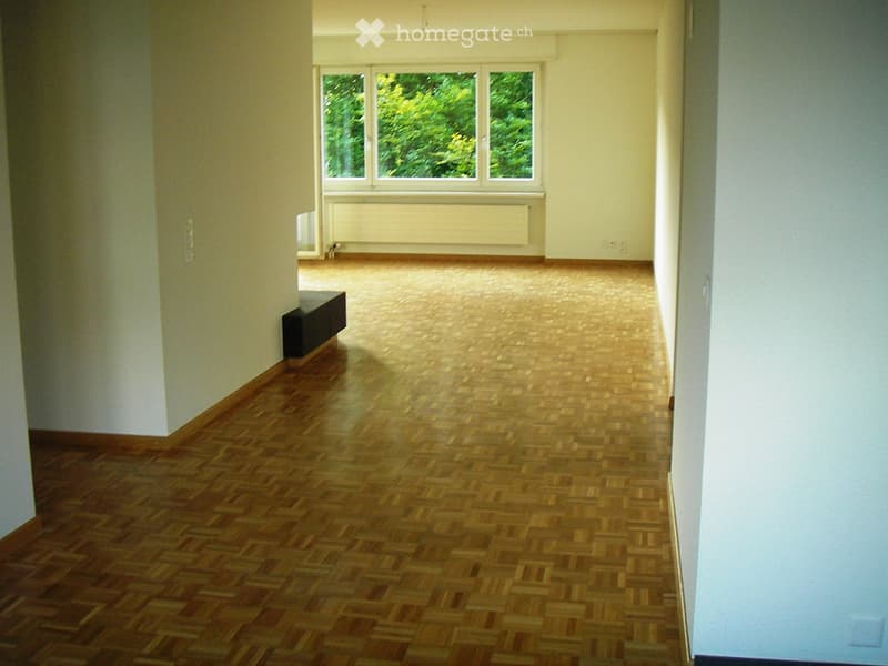 Empty room with wooden parquet flooring, white walls, and large windows with a view of greenery. No furniture or equipment present.