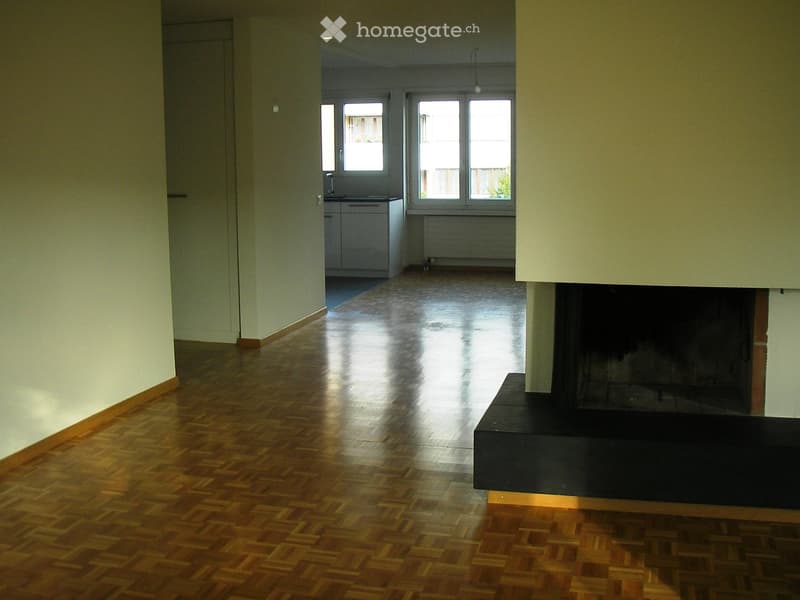 Empty hallway with wooden flooring, white walls, and a fireplace on the right side. Natural light streams in from windows at the far end.