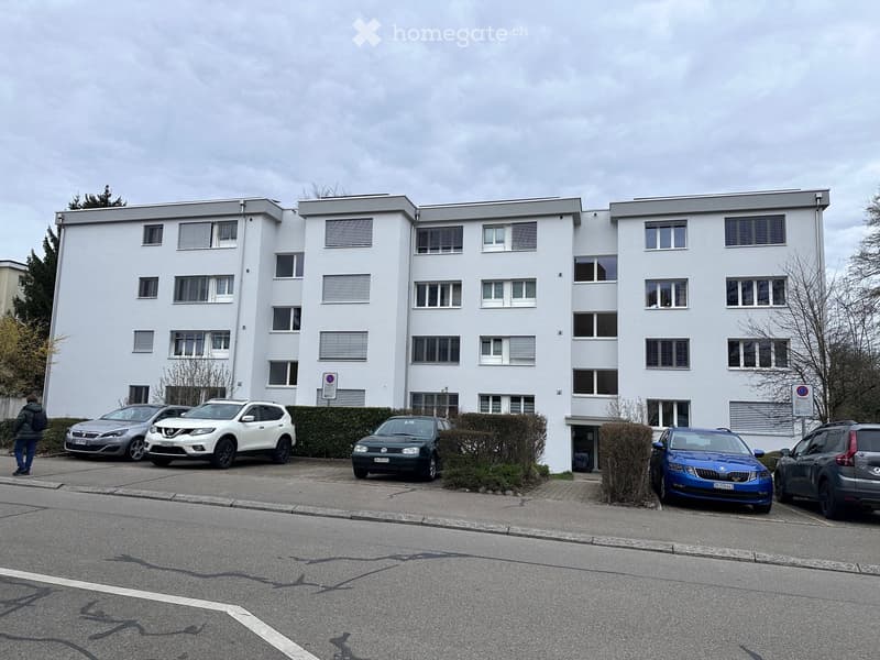 Modern multi-story apartment building with white facade, multiple windows, and several parked cars in front.