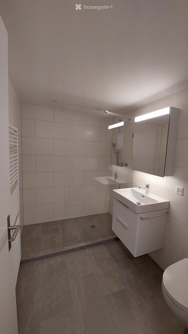 Modern bathroom with a white sink, mirror, toilet, and a glass-enclosed shower area. The walls are tiled in light beige, and the floor is made of large, light-colored tiles.