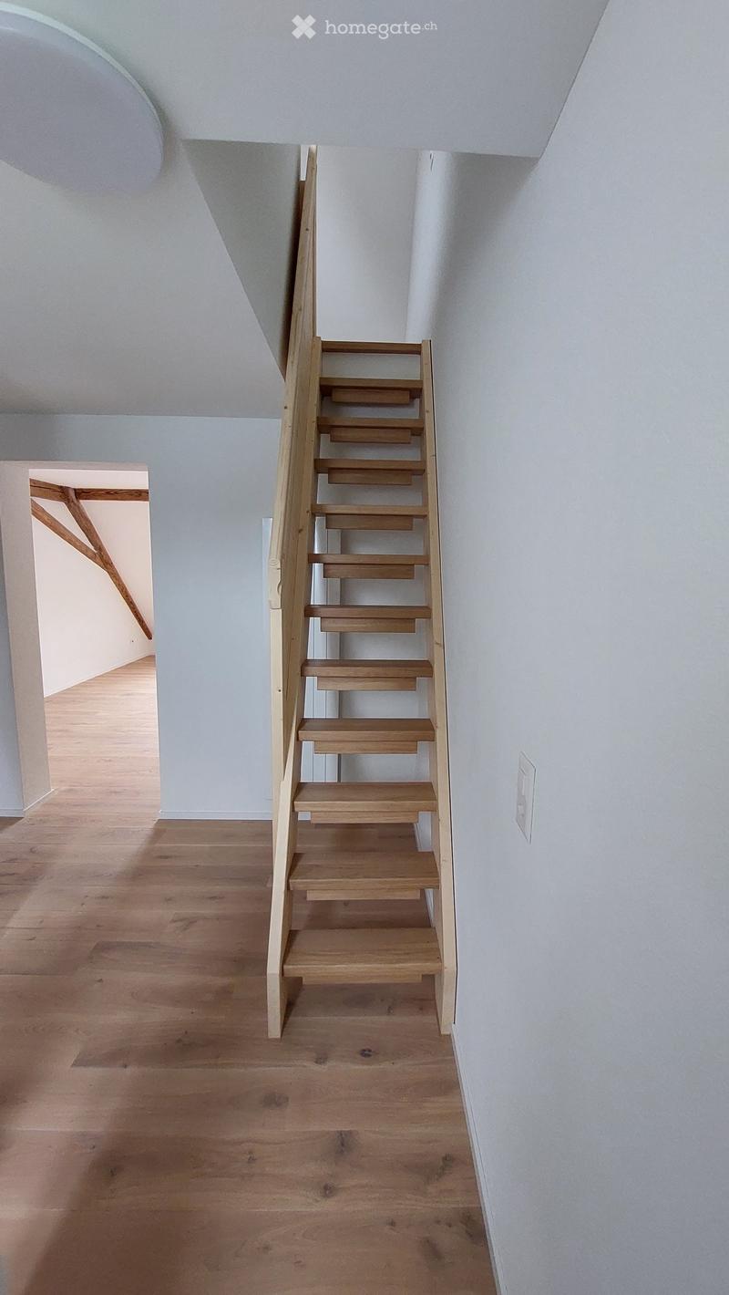 Wooden staircase with light-colored steps and railings, leading up from a wooden-floored hallway with white walls and a visible electrical outlet.