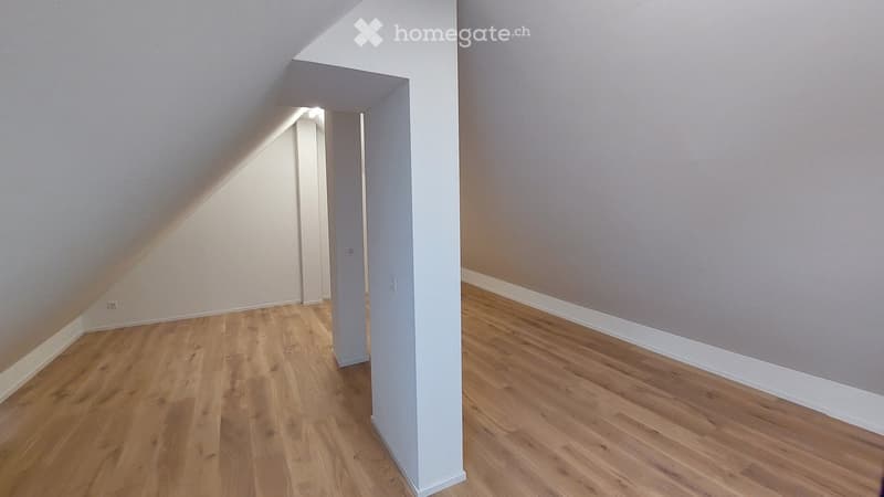 Empty attic room with wooden flooring, white walls, and a slanted ceiling. No furniture or equipment is present.