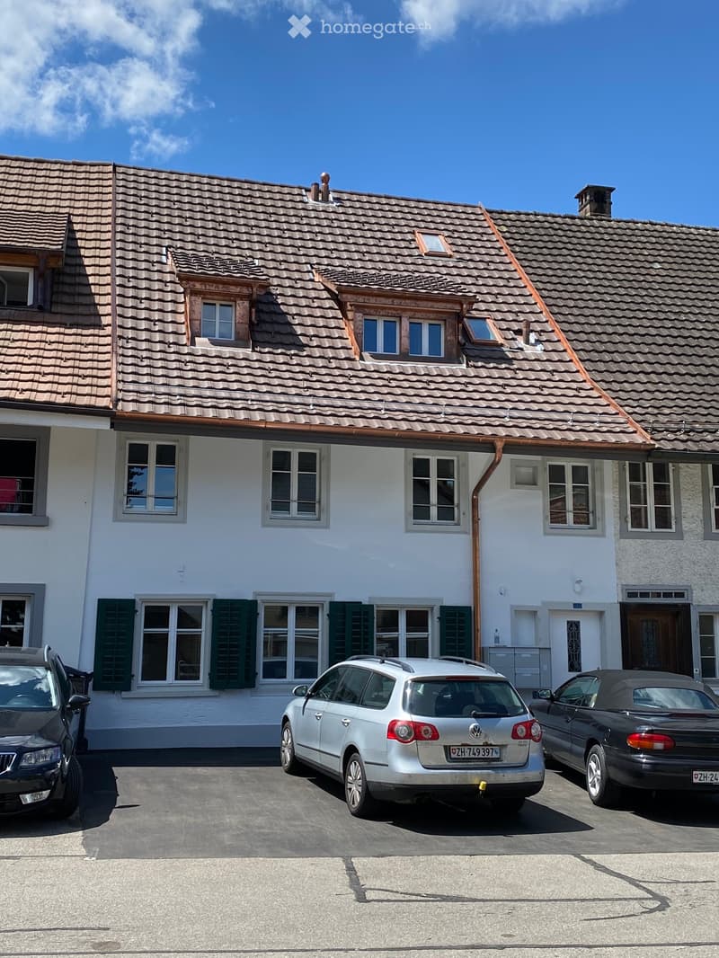 Two-story house with a tiled roof, white exterior, multiple windows with shutters, and a driveway with three parked cars.
