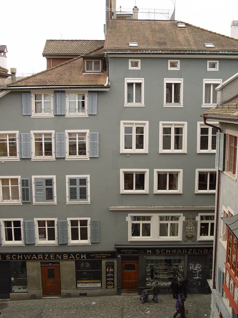 Four-story building with grey facade, multiple windows with shutters, and commercial storefronts on the ground floor.