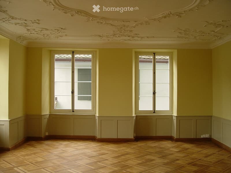 Empty room with wooden parquet flooring, light yellow walls, and two large windows with shutters. The ceiling features ornate plasterwork.