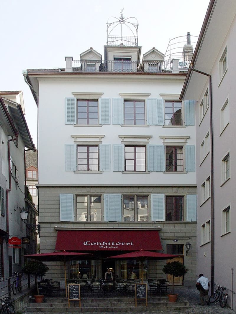 Three-story building with light-colored facade, multiple windows with shutters, and a red awning over the entrance. The ground floor features outdoor seating with tables and chairs under umbrellas.