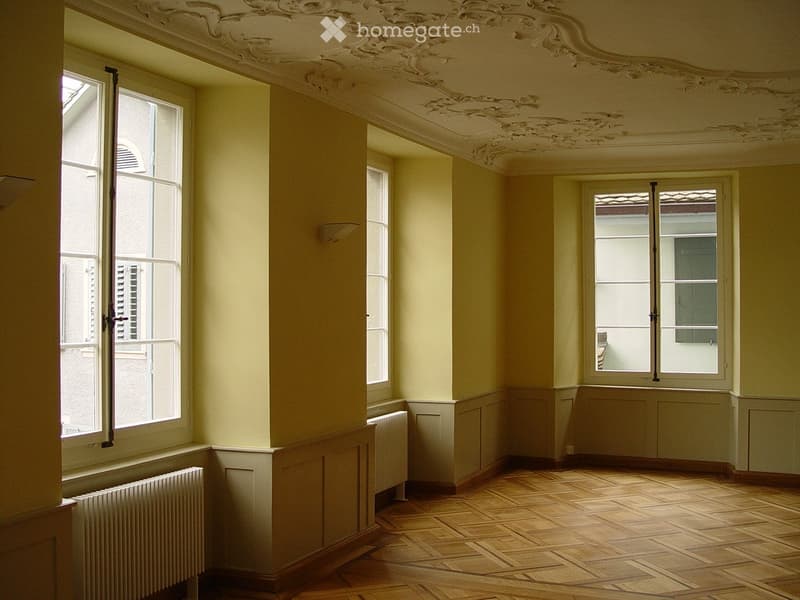 Empty room with wooden floor, large windows, radiators, and ornate ceiling design.
