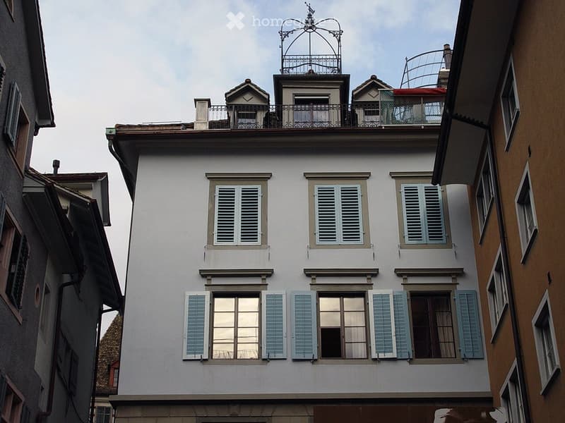 A multi-story residential building with white walls, multiple windows with shutters, and a decorative rooftop structure featuring a balcony and ornate metalwork.