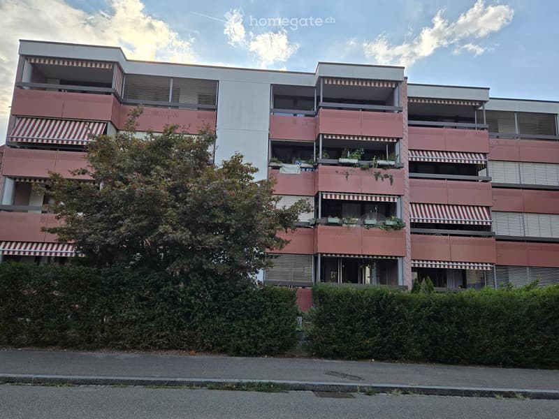 Multi-story residential building with pink and white facade, balconies, and greenery in front.
