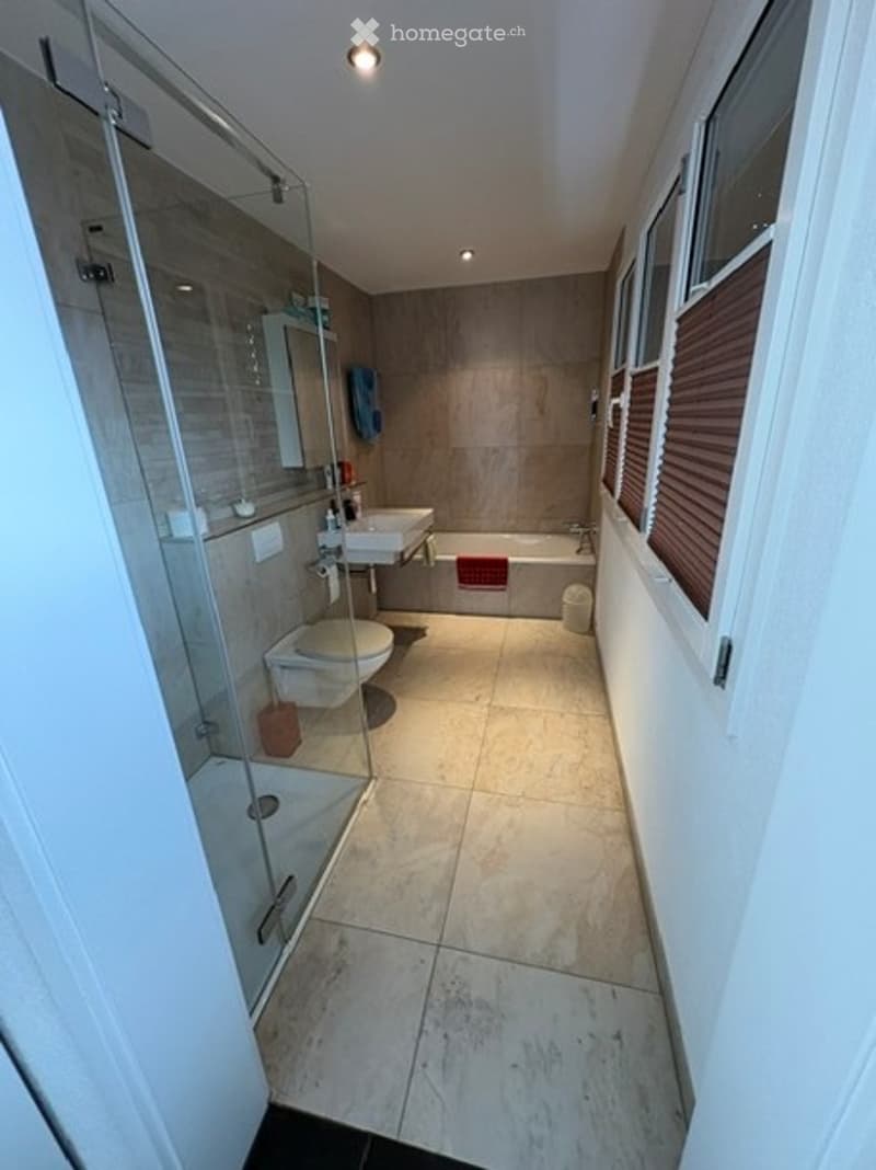 Modern bathroom with a glass-enclosed shower, toilet, sink, and bathtub. Light-colored tiles and a window with blinds provide natural light.