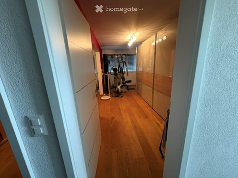 A narrow hallway with wooden flooring, white walls, and built-in mirrored cabinets on one side. A small exercise bike is visible in the adjacent room.