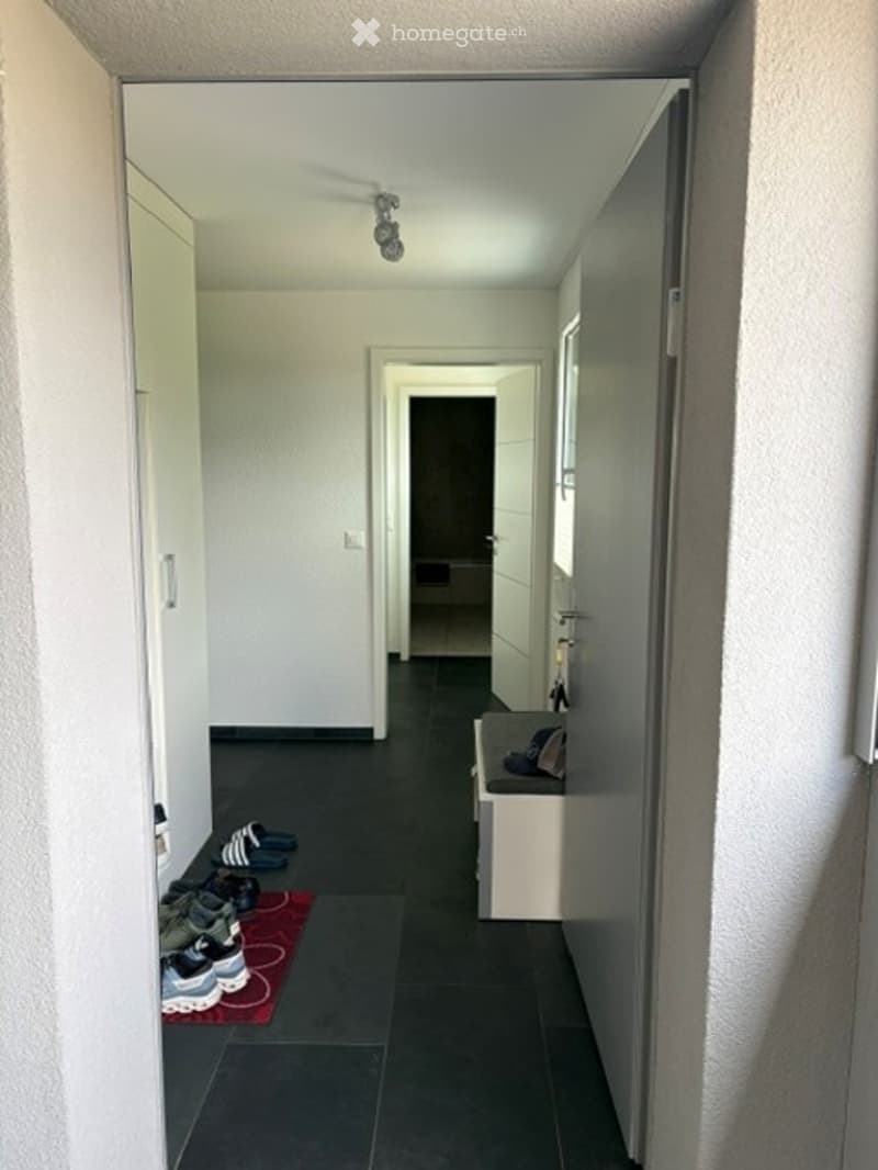 A narrow hallway with white walls, dark flooring, and a small bench with shoes placed on it. The hallway leads to another room with a visible doorway.