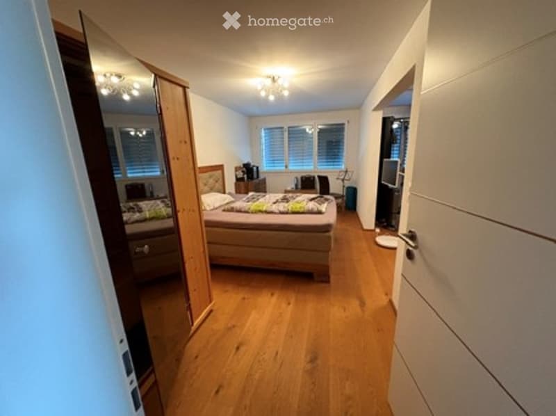 A furnished bedroom with a wooden floor, a bed with pillows and blankets, a nightstand, a mirror, a chair, and a small table with a lamp.