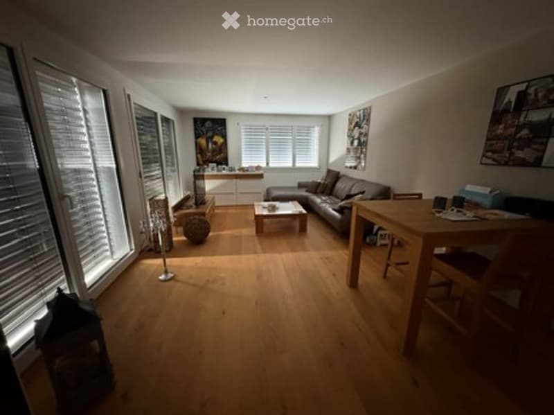 Bright living room with large windows, wooden flooring, a grey sectional sofa, a coffee table, a dining table with chairs, and a floor lamp. Artwork is displayed on the walls.