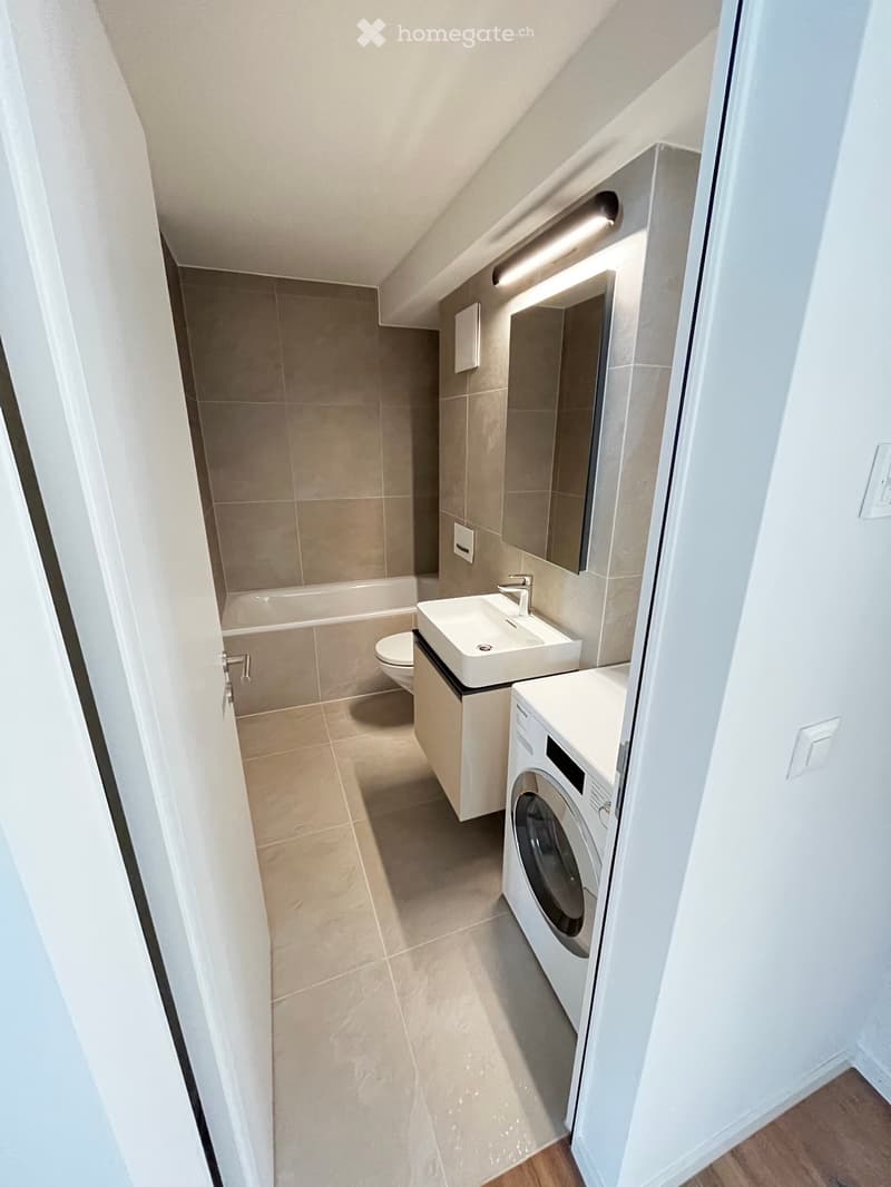 Modern bathroom with a sink, toilet, bathtub, and washing machine. Features a large mirror with built-in lighting and beige tiles.