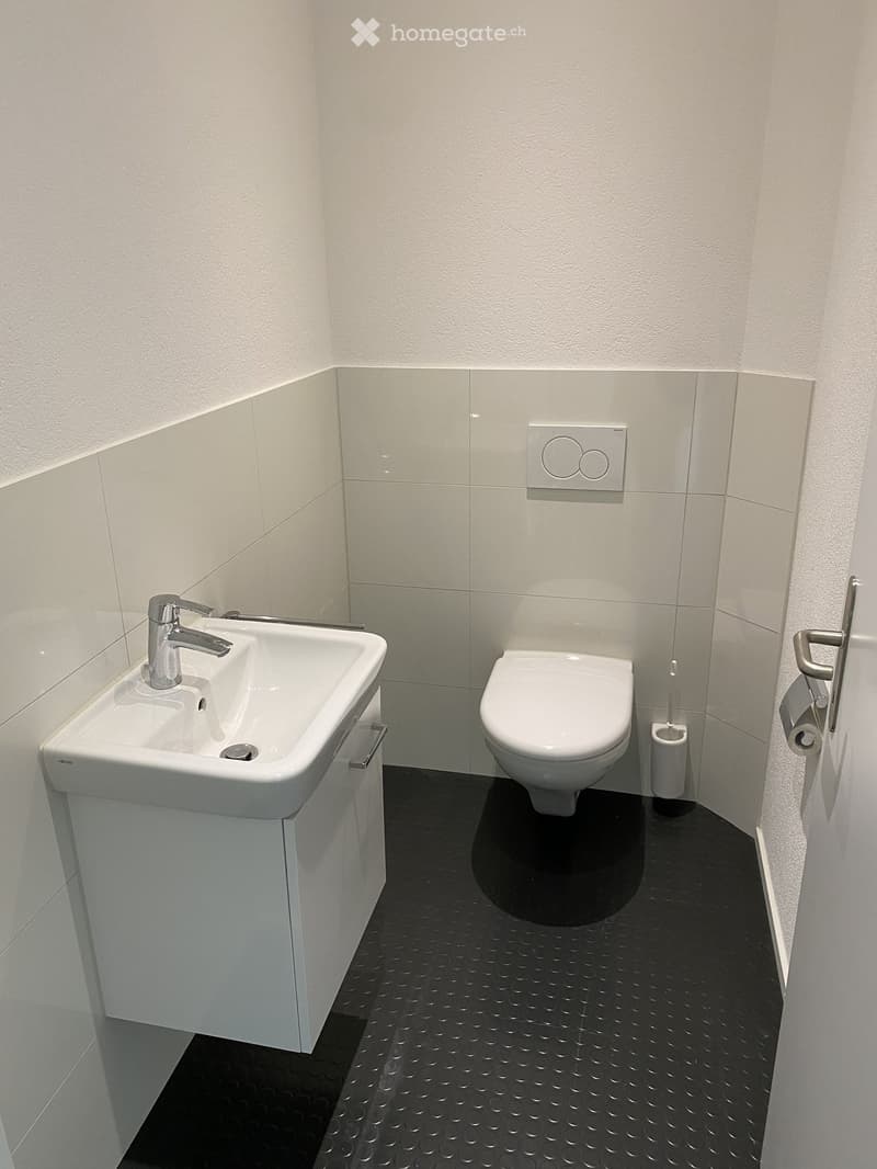 Modern bathroom with white tiles, a white sink with a chrome faucet, a toilet, and a toothbrush holder. The floor is black with a textured pattern.