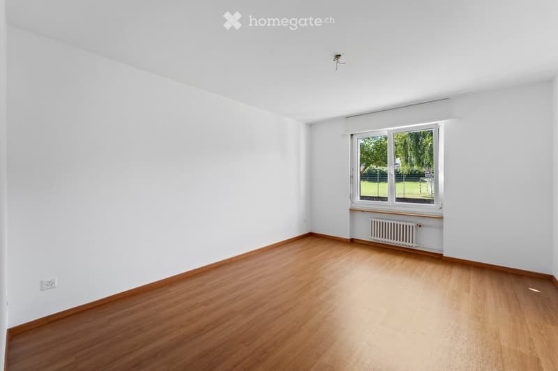Empty room with white walls, wooden floors, large window, radiator, electrical outlet
