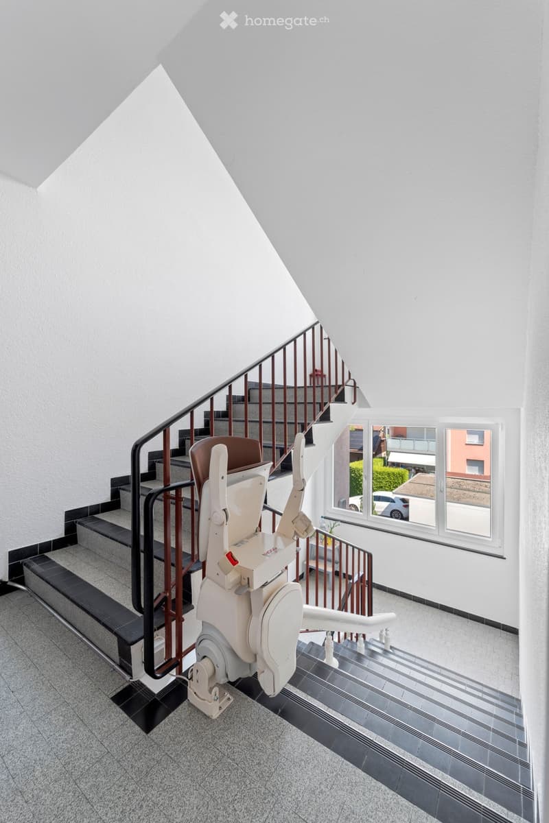 A flight of stairs with a lift chair in the middle, white walls, and a black tiled floor.