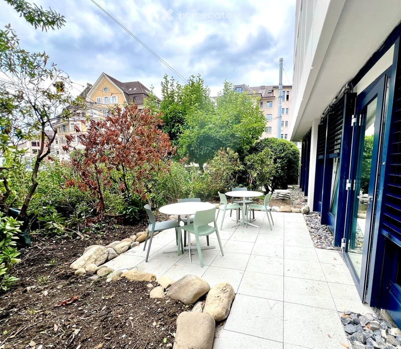 Outdoor patio area with a small seating arrangement, including a table and chairs, surrounded by greenery and rocks.