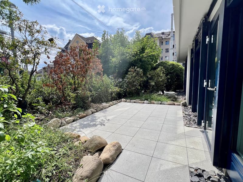 Garden area with a tiled pathway bordered by rocks and greenery, adjacent to a building entrance.