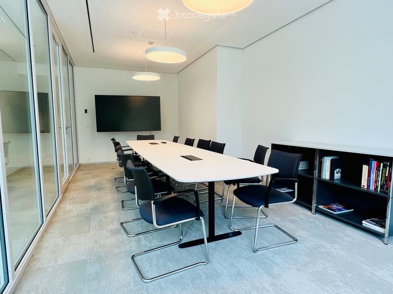 Modern conference room with a long white table, black chairs, a large screen on the wall, and a bookshelf with books and magazines.