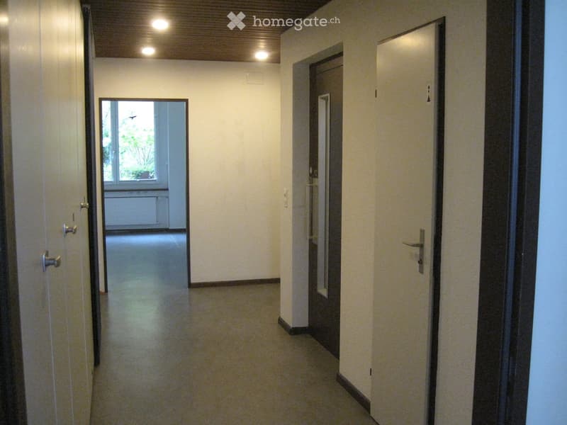 Empty hallway with beige walls, light-colored floor, and several doors. One door leads to a room with a window showing greenery outside.