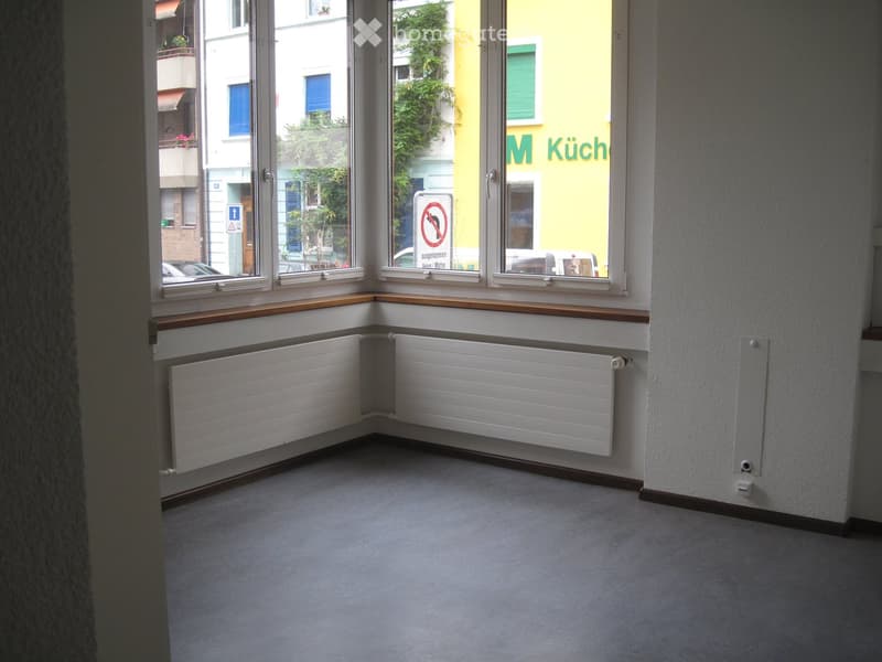 Empty room with large windows, radiators, and a wooden sill, showing an urban street view outside.