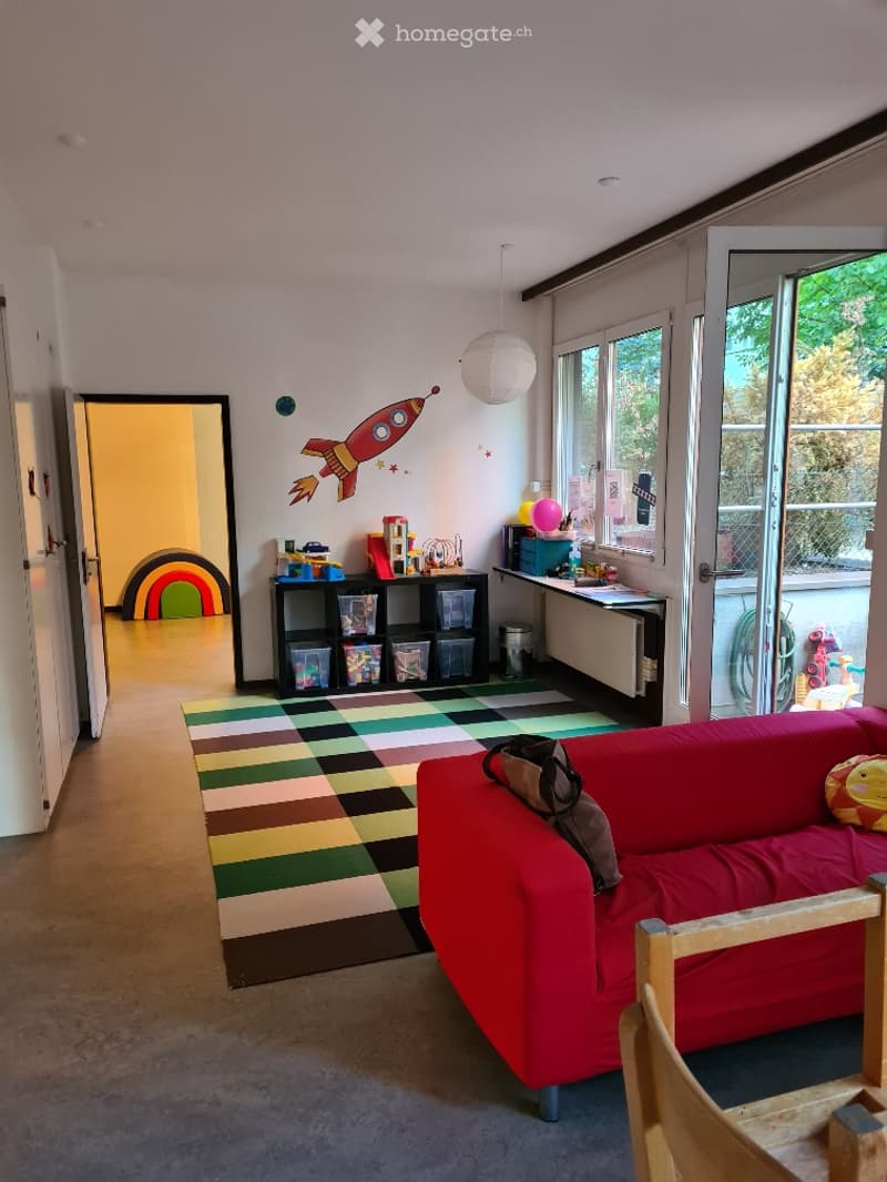 Bright, colorful living room with a red sofa, patterned rug, wooden shelf with storage bins, and a small table with toys and a balloon. A rocket ship mural adorns the wall, and a doorway leads to another room.