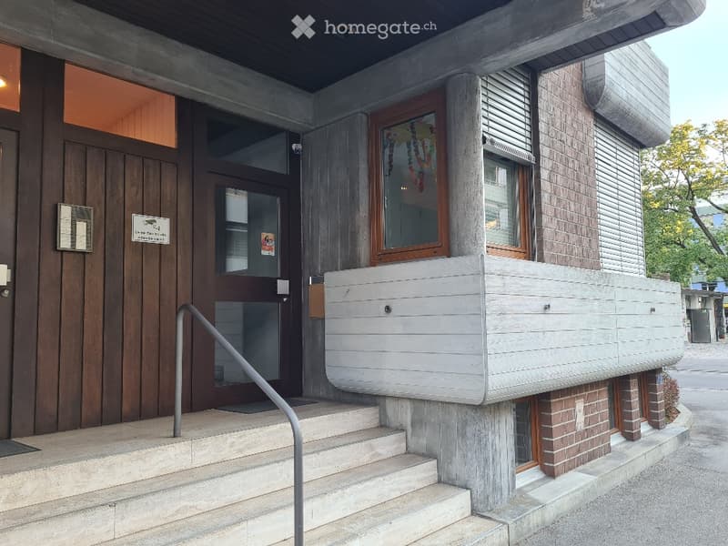 Modern building entrance with raised ground floor, concrete steps, metal railing, and a small balcony area with wooden paneling.