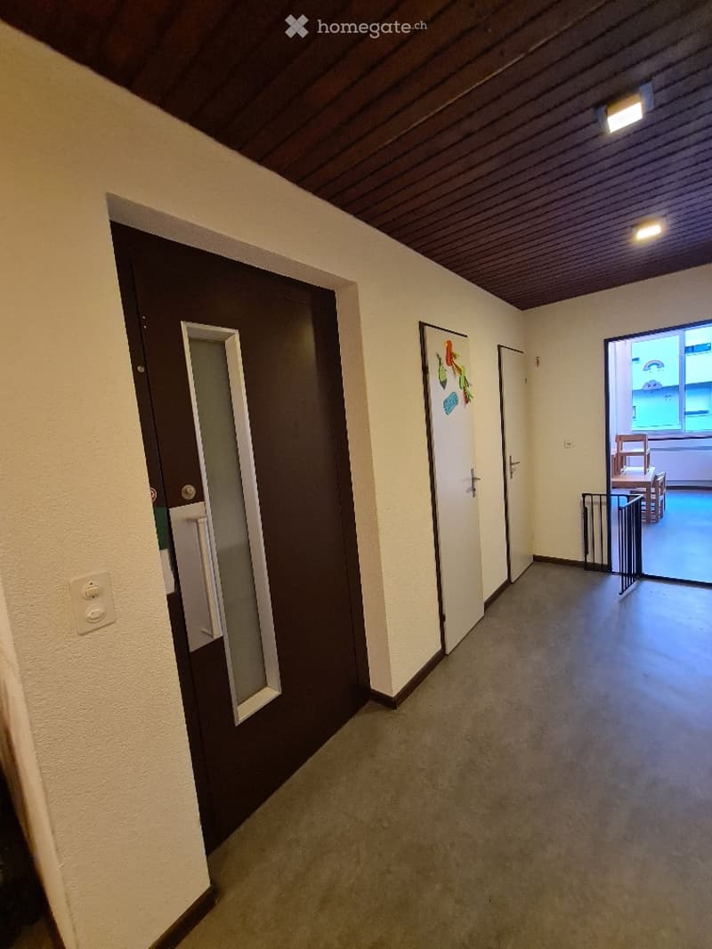 Empty hallway with beige walls, grey carpeted floor, and wooden ceiling. Features a dark brown door on the left, a white door with colorful stickers on the right, and an open doorway leading to another room with visible furniture.