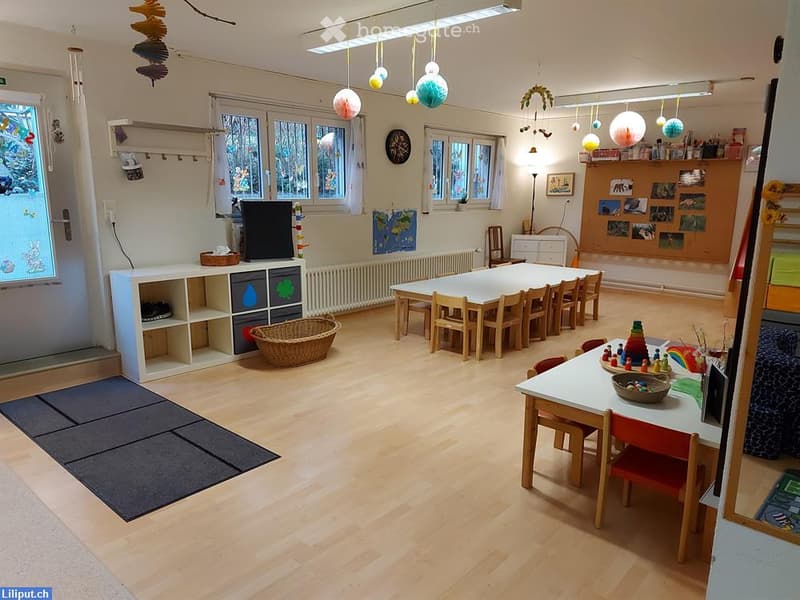 Brightly lit room with wooden flooring, large windows, and colorful decorations. Features a long table with chairs, a small shelf unit, a wicker basket, and various toys and educational materials.