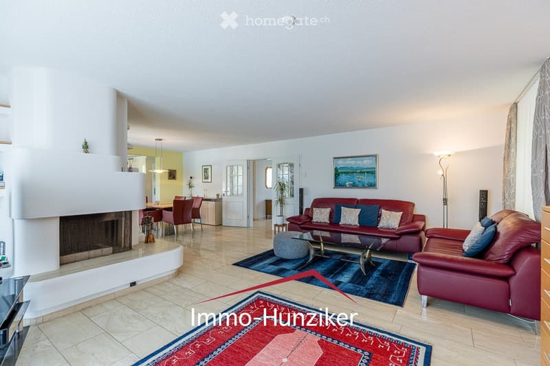 Modern living room with a fireplace, red leather sofa, armchair, coffee table, ottoman, dining area with table and chairs, floor lamp, and artwork on the wall.
