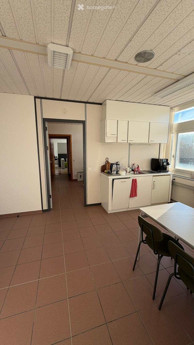 An empty room with brown tiles and white walls. It features a kitchen area with appliances like a coffee machine, kettle, and microwave. The room also has a table, chairs, and cabinets.