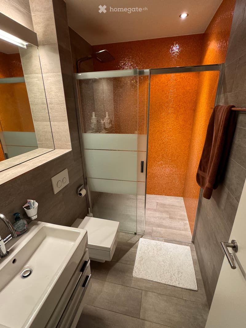 Modern bathroom with a white sink, toilet, glass-enclosed shower, and a towel hanging on a rack. The walls are tiled in neutral tones, and the floor is covered with beige tiles.