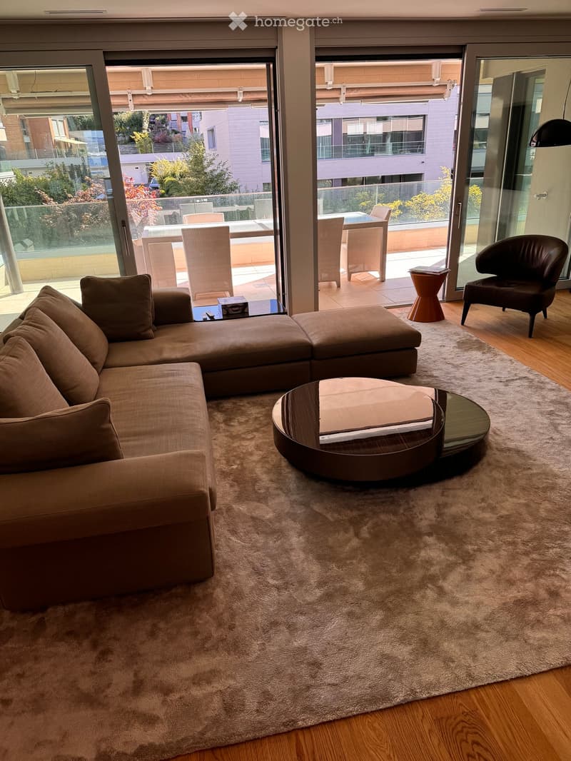 Modern living room with a large sectional sofa, coffee table, and a view of a balcony with outdoor furniture.
