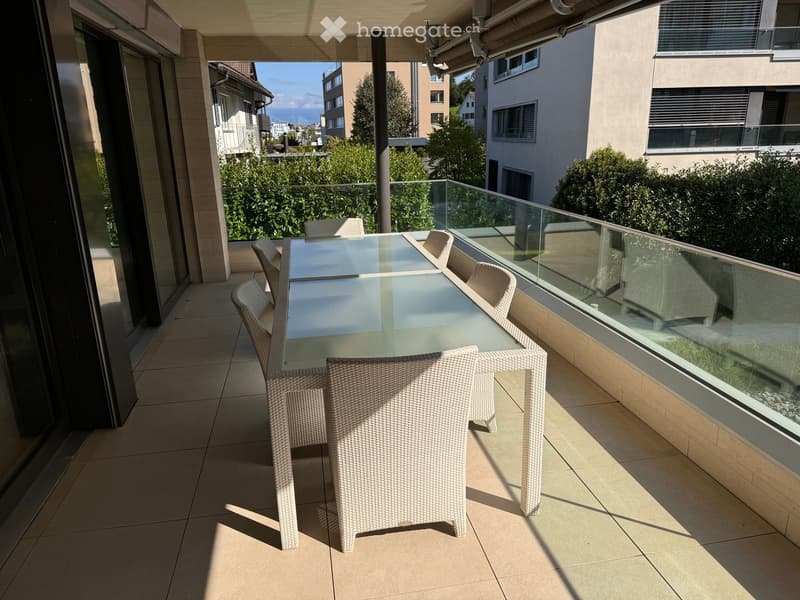 Balcony with a glass table and wicker chairs, overlooking a residential area with greenery and buildings.