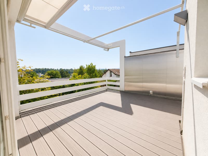 Balcony with wooden decking, white railing, retractable awning, and scenic view of greenery and distant buildings.
