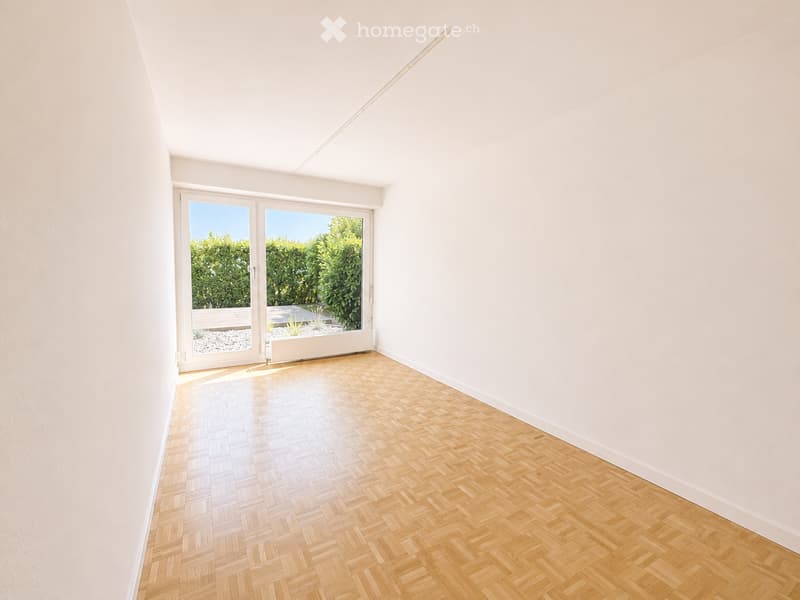 Empty room with light wooden herringbone flooring, white walls, and large glass doors leading to a garden area.