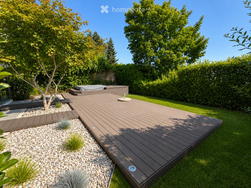 A modern wooden deck with integrated hot tub, surrounded by neatly trimmed greenery and a gravel pathway.