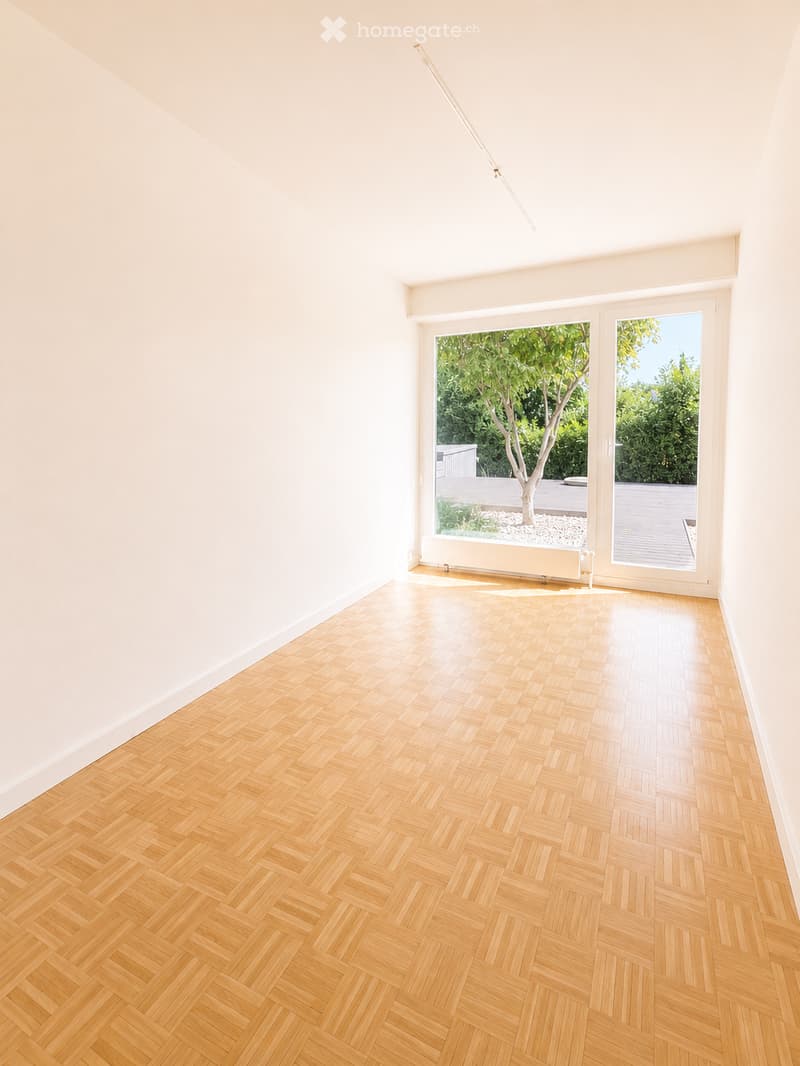 Empty room with light wooden herringbone flooring and white walls, featuring large glass doors leading to an outdoor area with a tree and patio.