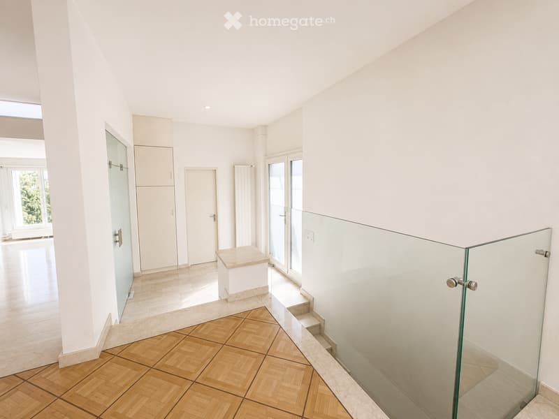 Modern hallway with white walls, wooden herringbone flooring, glass railing, and built-in storage unit.