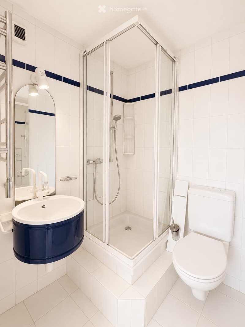 Modern bathroom with white tiles, a blue-accented sink, a glass-enclosed shower, and a toilet. Features include a large mirror with integrated lighting and a sleek, contemporary design.
